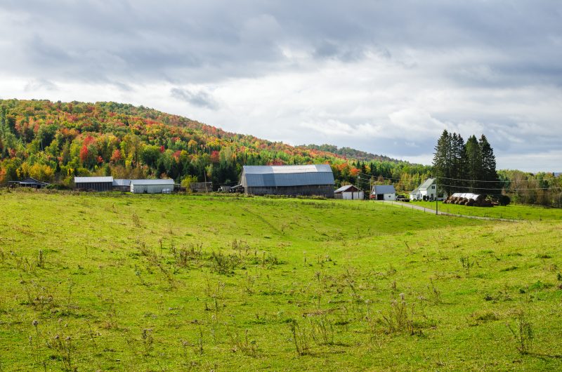 Beautiful rural landscape with farm buildings at the foot of a wooded hill and storm sky. Autumn Colours. New Brunswick, Canada.